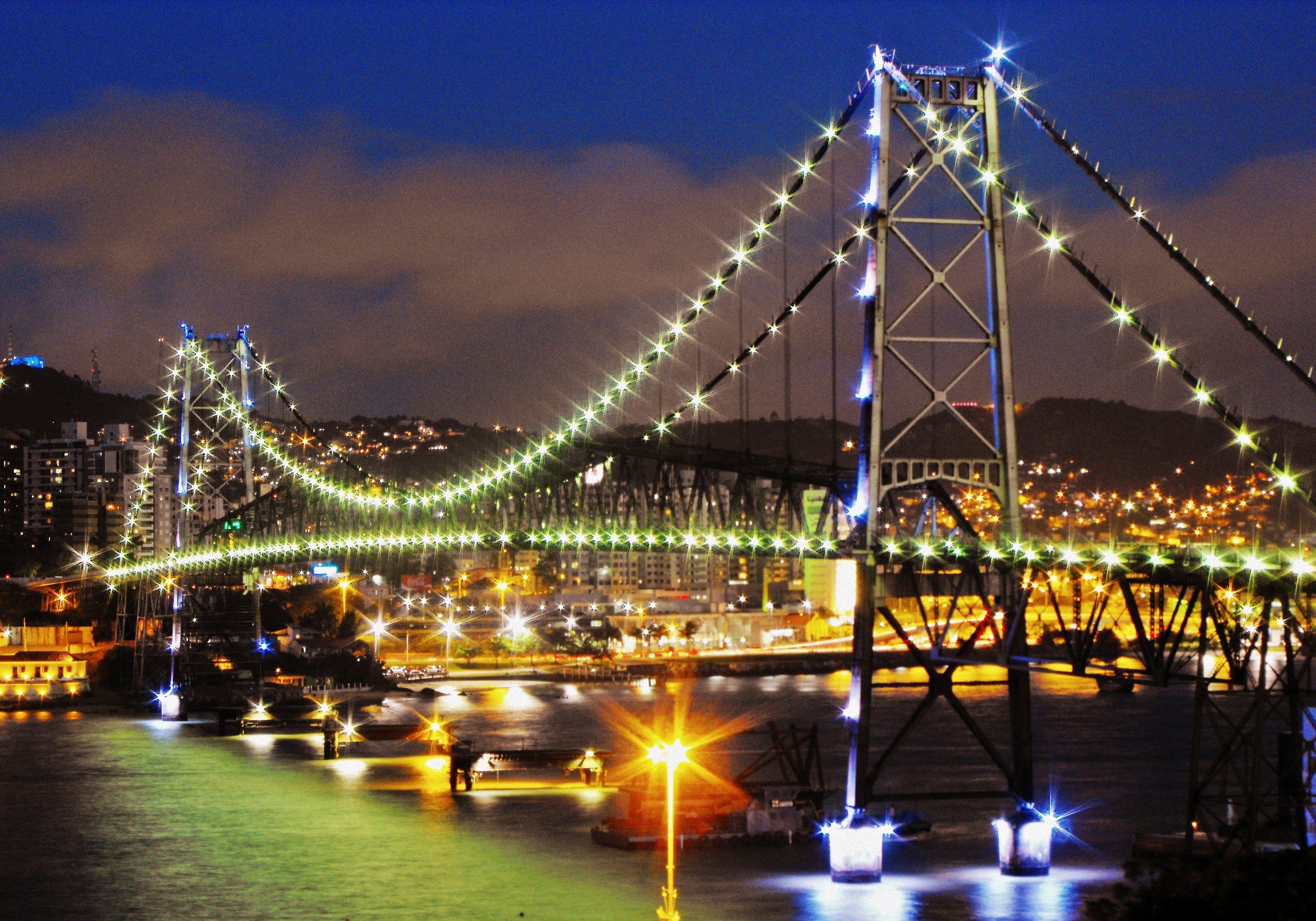 A Ponte Hercílio Luz iluminada à noite, com os cabos marcados por luzes e o reflexo no canal entre a ilha e o continente