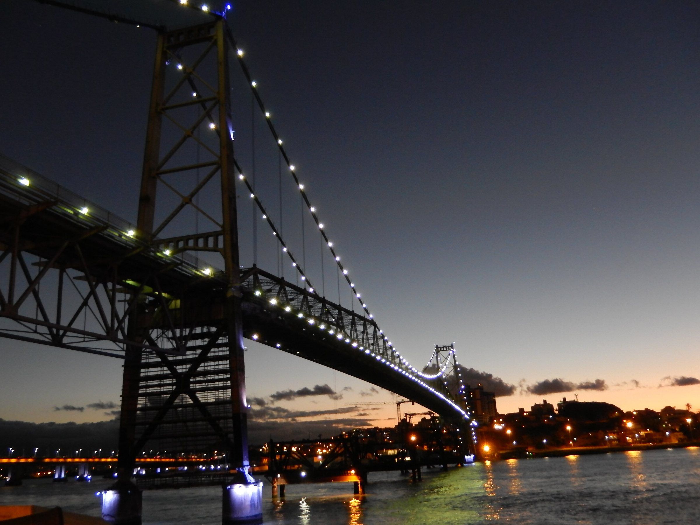 A torre e o tabuleiro da Ponte Hercílio Luz ao entardecer, com as luzes acesas e o céu alaranjado