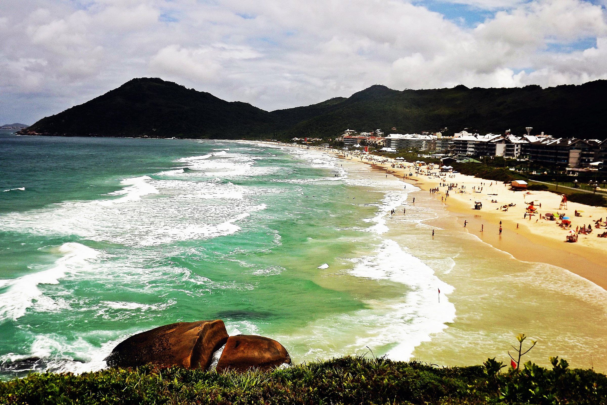 A Praia Brava vista do costão, com as ondas brancas, o mar verde e os morros