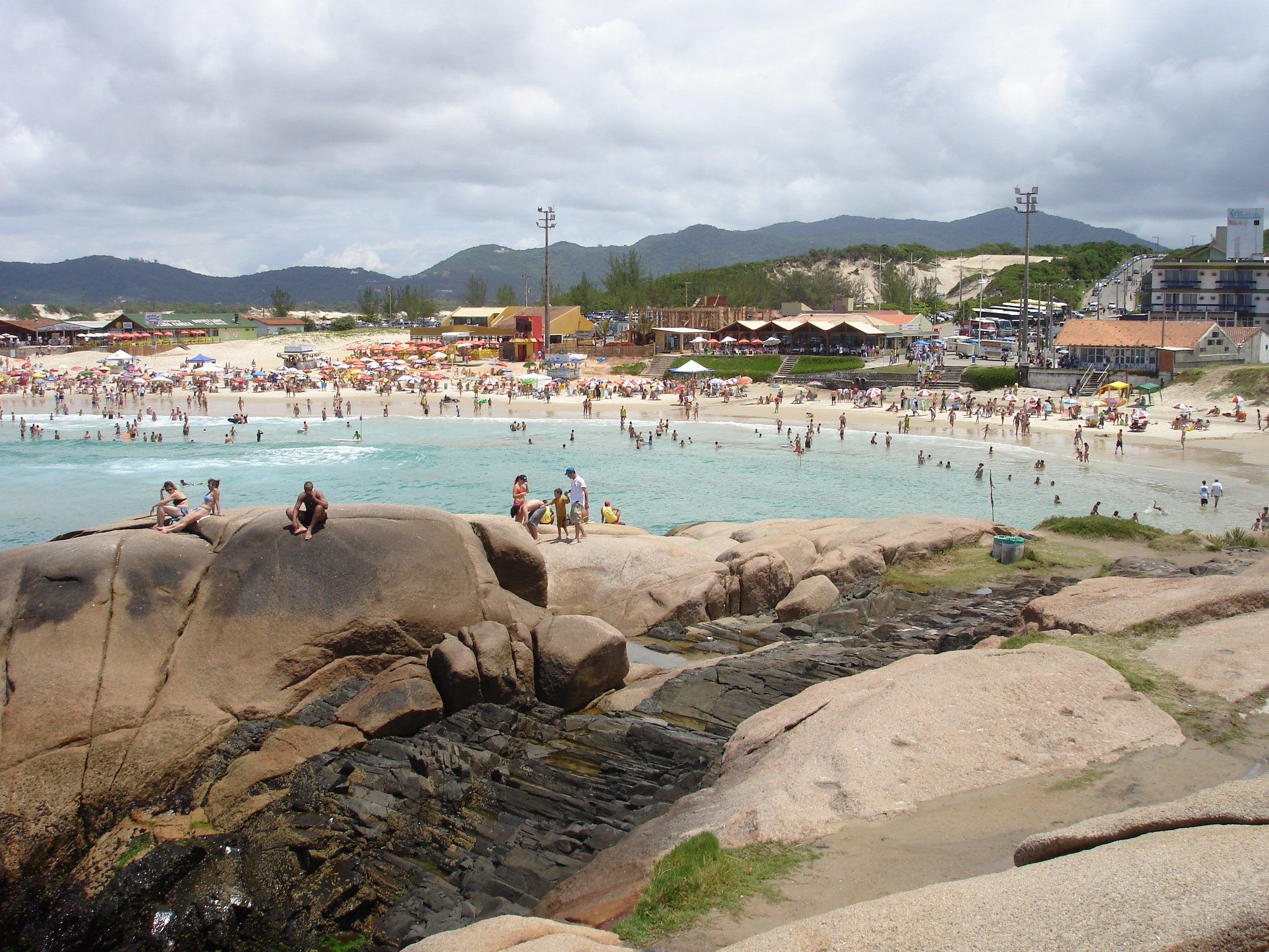 A Praia da Joaquina cheia no verão, vista das pedras do costão, com o mar verde e as dunas ao fundo