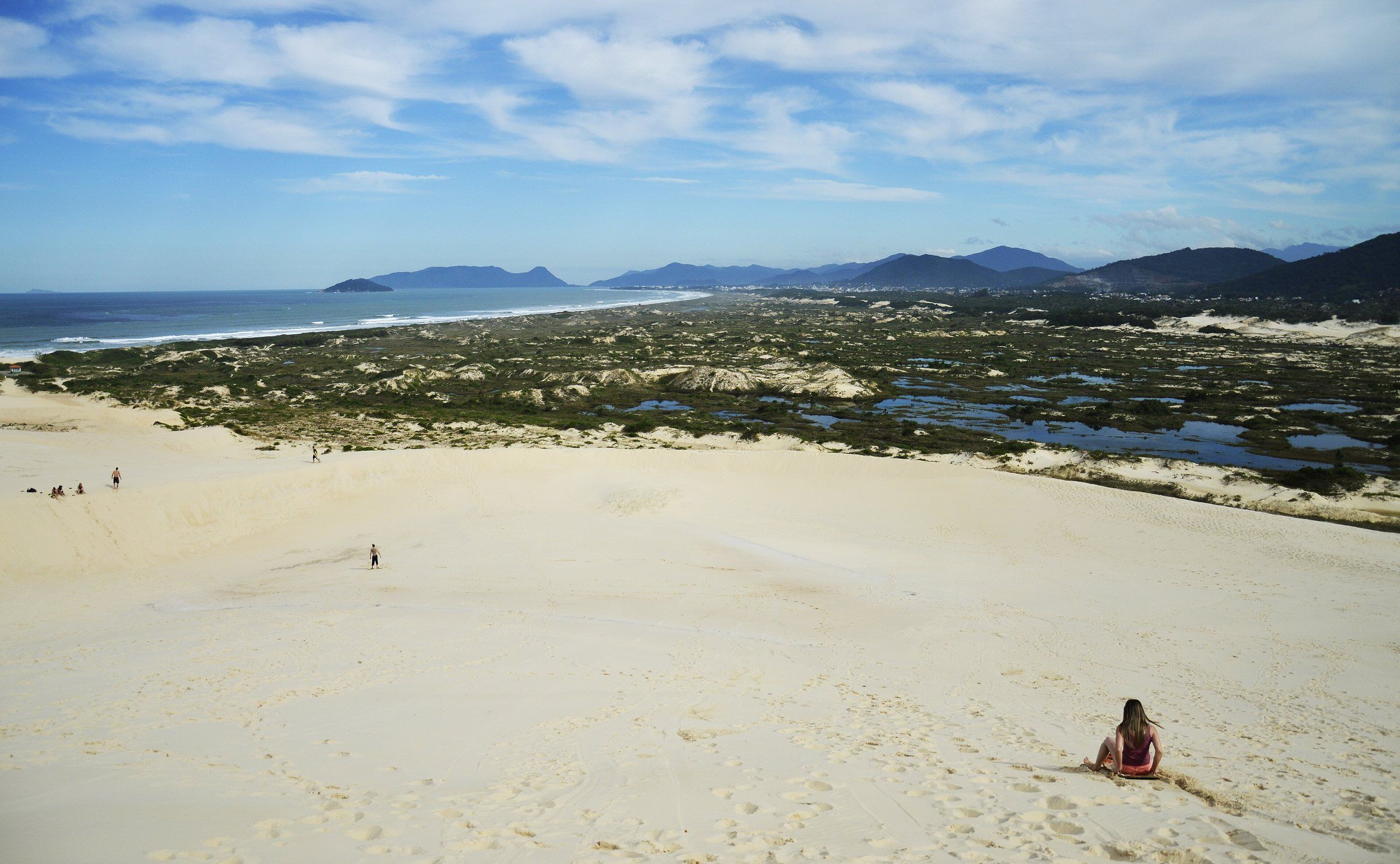 Pessoas descendo as dunas da Lagoa da Conceição de prancha (sandboard), com a Praia da Joaquina ao fundo